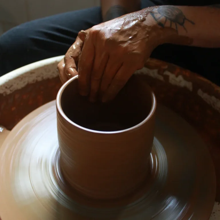Hands shaping a clay cylinder on the spinning pottery wheel at Klei